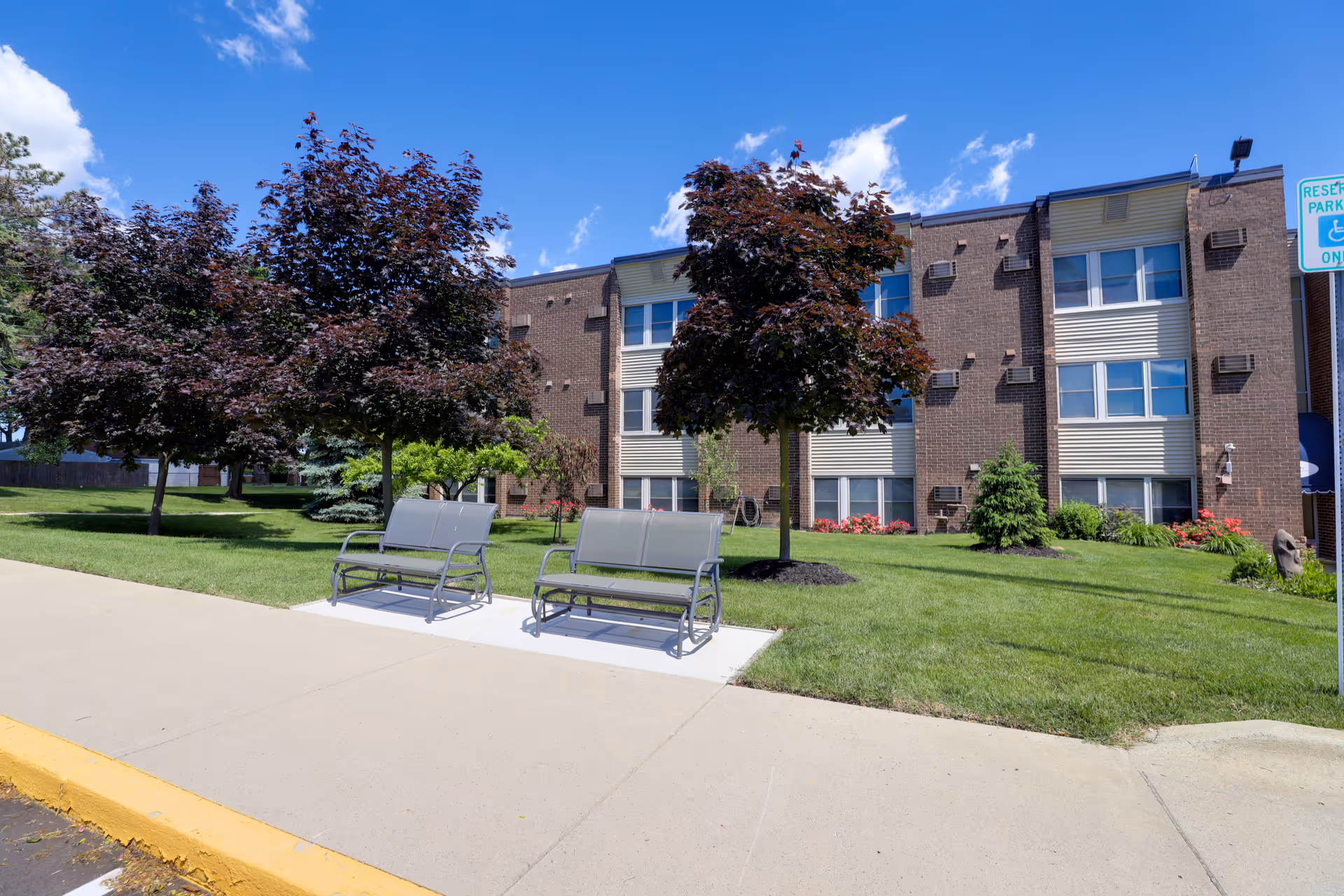 Front exterior of a three-story brick senior living building with two benches on a concrete pad, trees, and a grassy lawn under a blue sky.