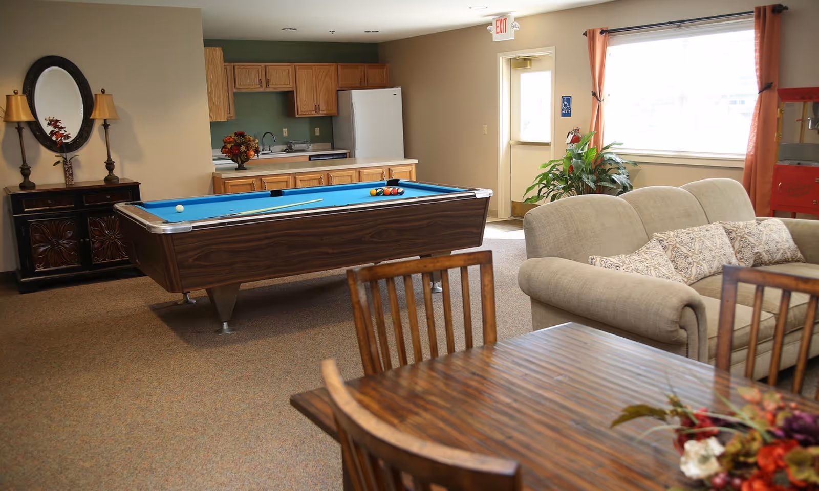 A cozy common area featuring a pool table with a blue felt top, a beige couch with patterned pillows, a wooden dining table with chairs, a small kitchenette with wooden cabinets and a white refrigerator, a decorative mirror with two lamps on a dark wooden cabinet, a large window with orange curtains, and a red popcorn machine near the window.