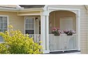 Front porch of a beige senior living building with white railing and potted flowers.