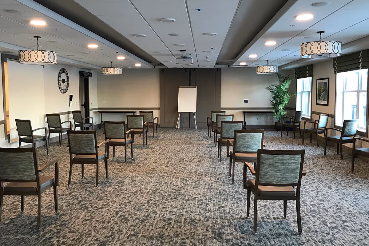 A spacious meeting or activity room with multiple chairs arranged in rows facing a blank flip chart on an easel. The room has carpeted flooring, modern ceiling lights, large windows with green blinds, a large wall clock, and a potted plant in the corner.