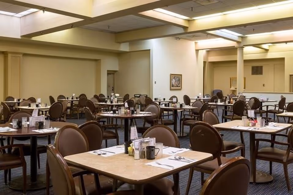 A spacious dining room with multiple round and square tables set with placemats, utensils, cups, and condiments. The room has beige walls, a carpeted floor, and overhead lighting with exposed beams. Several brown cushioned chairs are arranged around the tables.