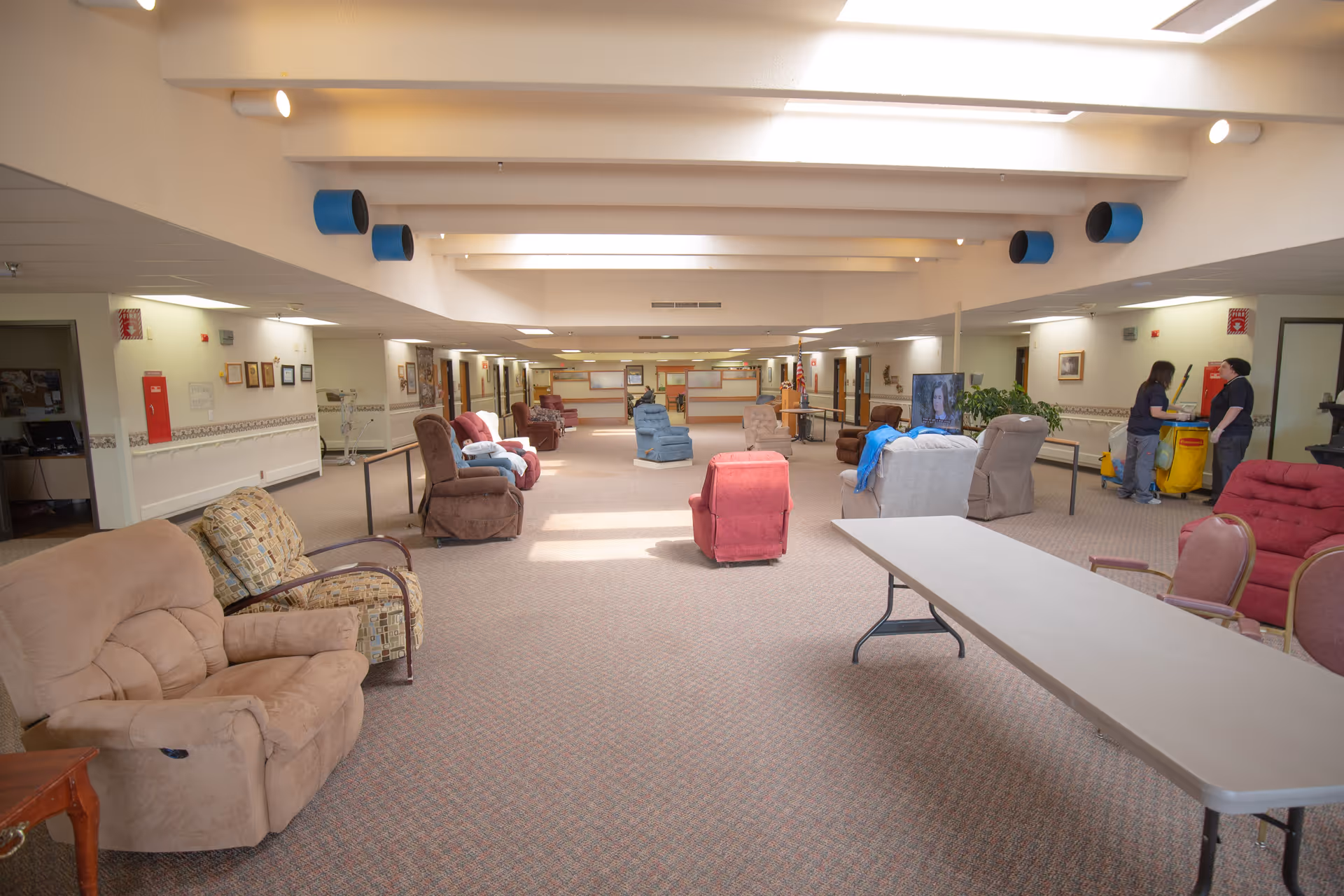 Spacious communal lounge with rows of recliners, a folding table, and two staff by a cleaning cart.