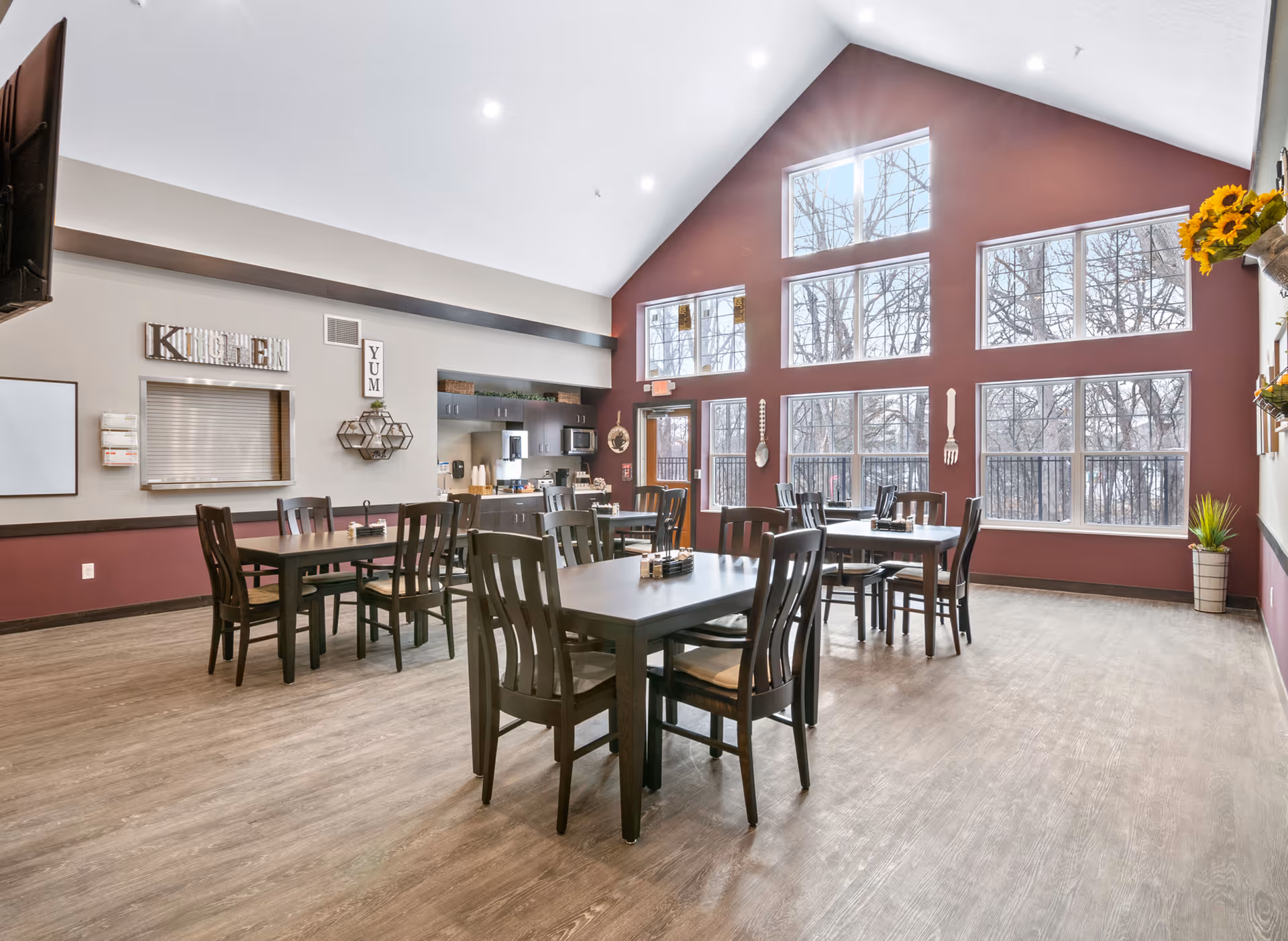A bright dining room with several dark wooden tables and chairs arranged neatly. The room features large windows on a maroon accent wall, allowing natural light to fill the space and providing a view of leafless trees outside. The opposite wall has a small kitchen serving window with decorative signs reading 'KITCHEN' and 'YUM'. The floor is wood-patterned, and the ceiling is vaulted with recessed lighting. There are decorative items such as large spoon and fork wall hangings and a small plant in the corner.