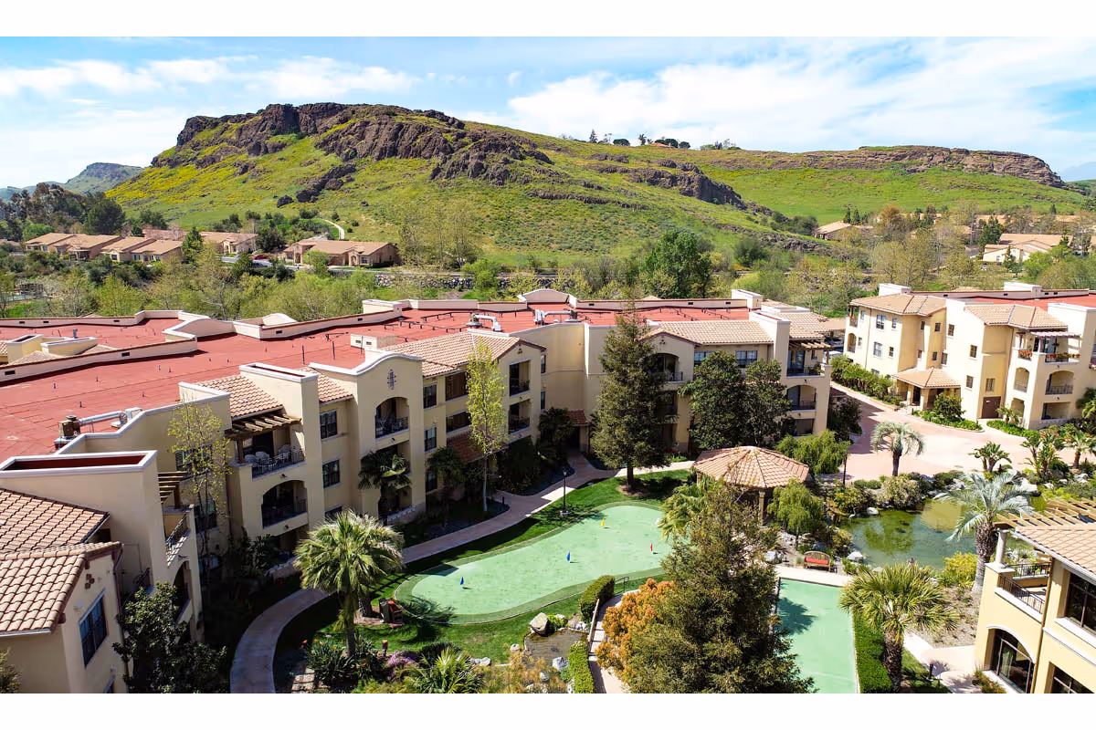 Aerial view of University Village Thousand Oaks senior living facility with multiple beige buildings featuring red-tiled roofs, surrounded by landscaped gardens, palm trees, a small pond, and a putting green. In the background, there is a large green hill under a partly cloudy sky.