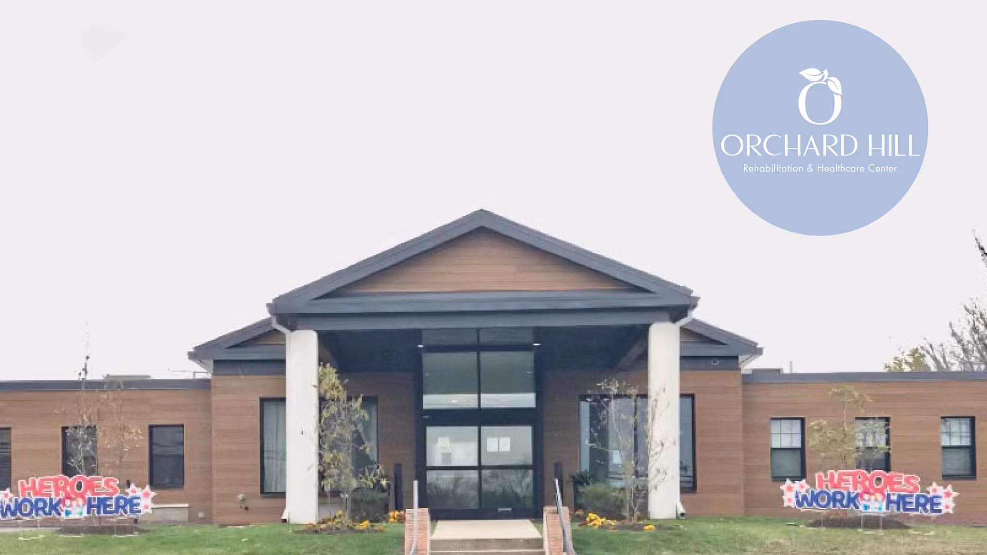 Front exterior view of Orchard Hill Rehabilitation & Healthcare Center building with a covered entrance supported by white columns, brown siding, and windows. There are two signs on the lawn that say 'HEROES WORK HERE' in red and blue letters with stars. The facility logo is visible in the top right corner.