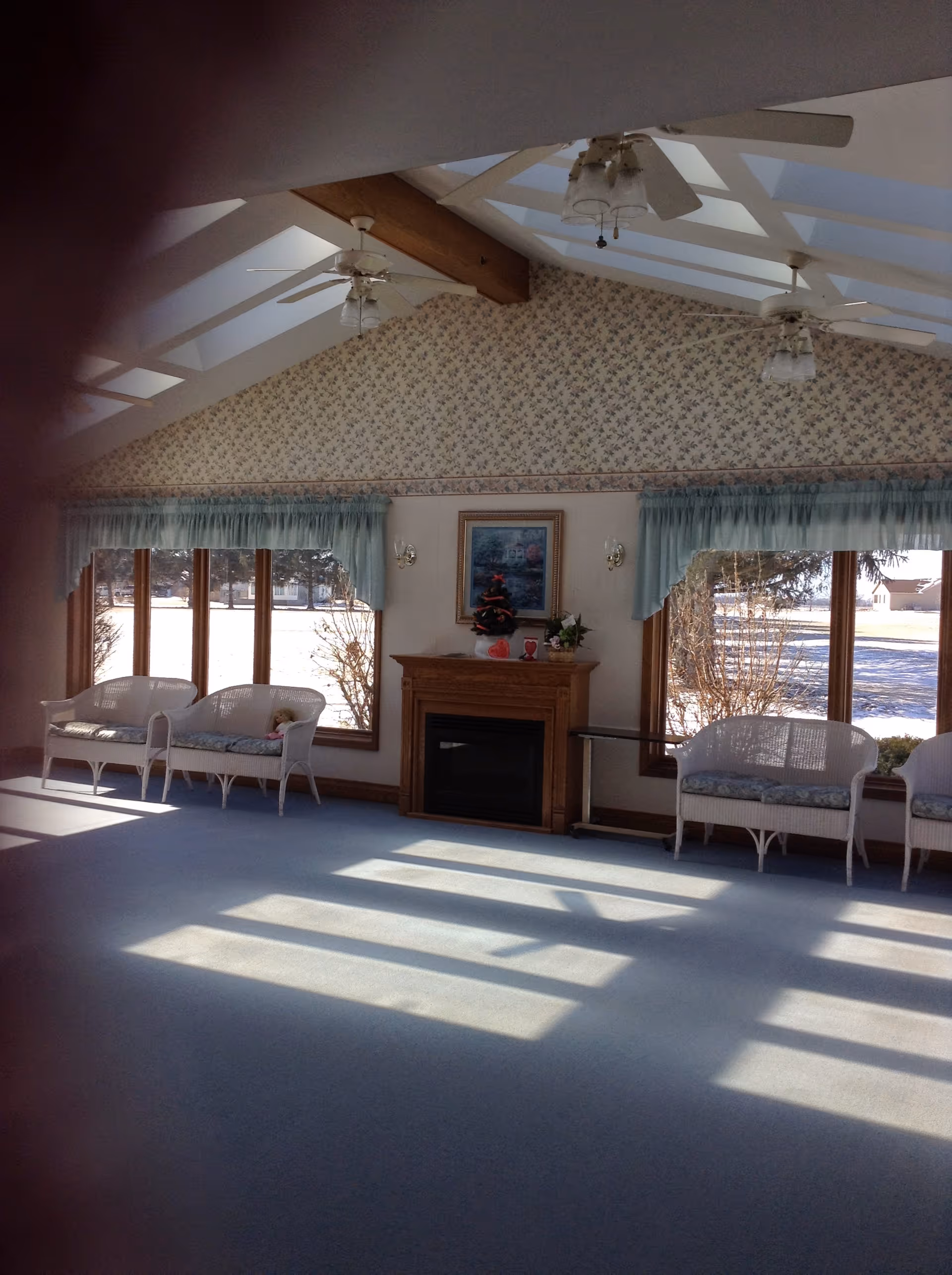 A bright, spacious room with large windows letting in sunlight, white wicker chairs with cushions arranged along the walls, a wooden fireplace with a small decorated Christmas tree on top, floral wallpaper on the upper wall, and ceiling fans with lights hanging from a vaulted ceiling with exposed beams.