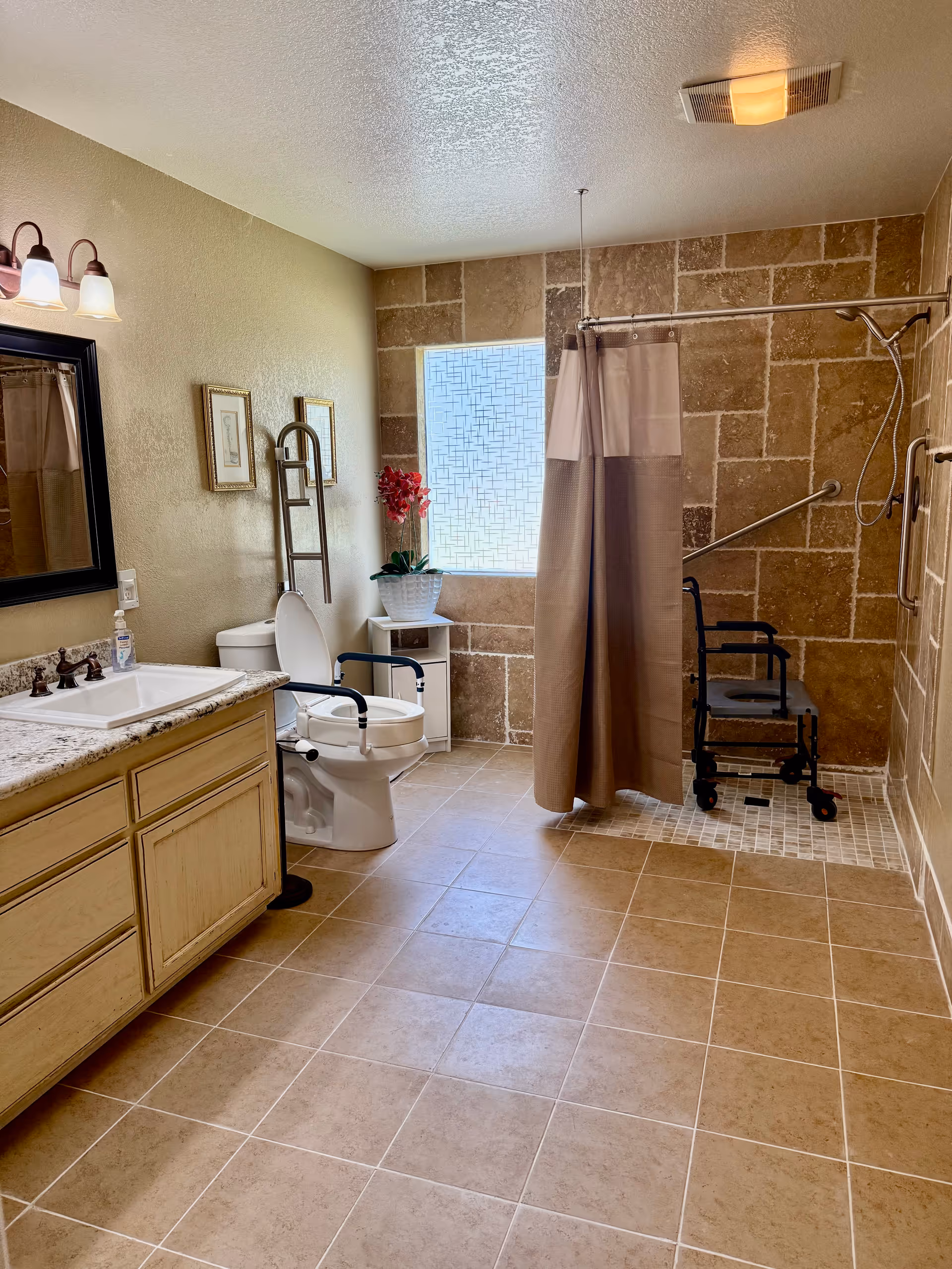 Spacious bathroom with beige tiled floor and walls, featuring a sink with granite countertop and wooden cabinets, a toilet with safety rails, a window with frosted glass, a shower area with a curtain and a shower chair, and decorative elements including framed pictures and a potted plant.