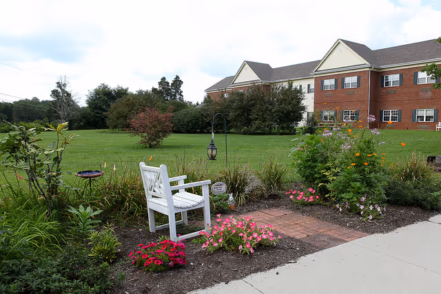 A white garden bench amid colorful flower beds and a lamp on a paved path with a brick-faced assisted living building and lawn in the background.