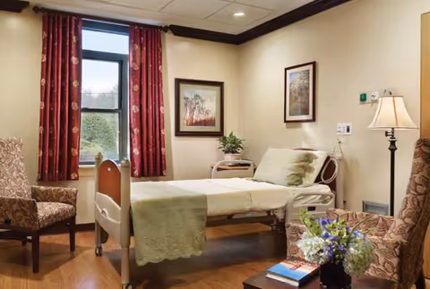 A cozy hospital-style bedroom with a bed, seating, window, and framed art.