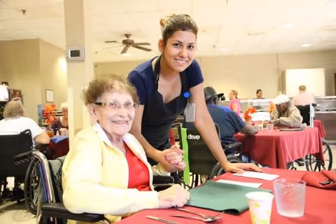 A smiling elderly woman in a wheelchair wearing a yellow jacket and red shirt sits at a table with a young female caregiver standing beside her, both looking at the camera. The setting is a dining area with other elderly people seated at tables covered with red tablecloths.