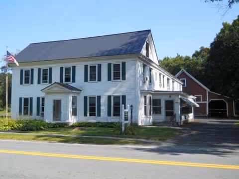 White two-story residence with shutters, a porch, flag, and a red barn beside.