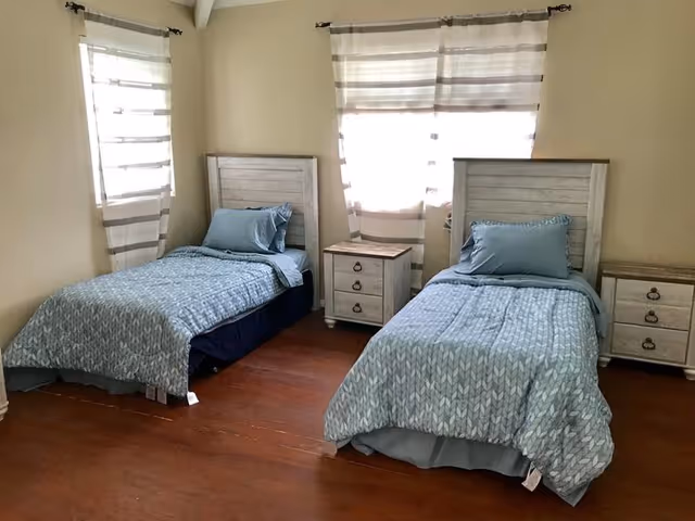 Two twin beds with light blue bedding and matching wooden headboards and nightstands in a sunlit bedroom with hardwood floors and striped curtains.