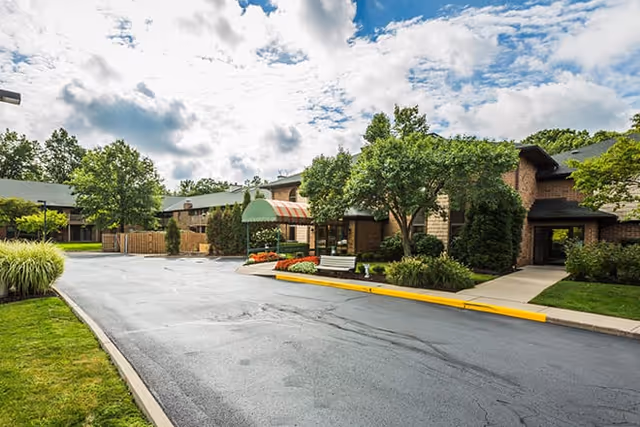 Exterior view of Brookdale Gardens at Westlake showing a paved driveway, landscaped greenery, trees, and a building entrance with a green and red striped awning under a partly cloudy sky.