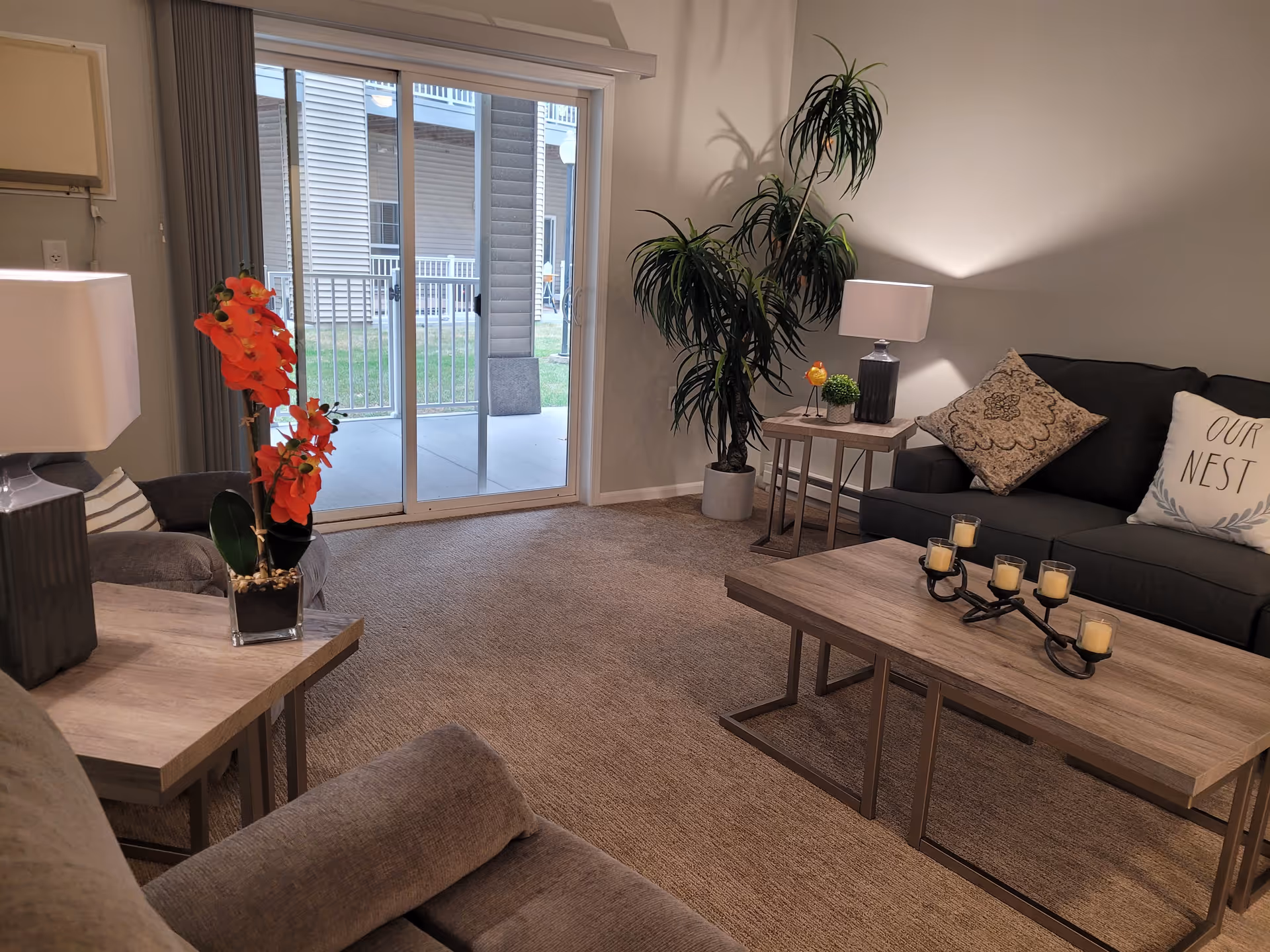 A cozy living room with a dark gray sofa adorned with decorative pillows, including one that says 'OUR NEST'. There is a wooden coffee table with a candle holder featuring five candles, a side table with a lamp, and a large potted plant in the corner. A sliding glass door leads to an outdoor patio area with a view of neighboring buildings. The room is carpeted and has neutral-colored walls.