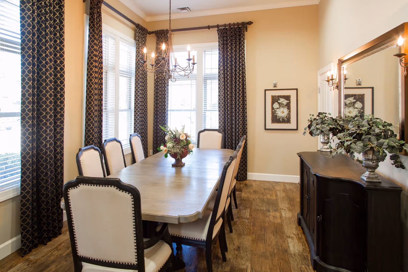 A well-lit dining room with a long wooden table surrounded by eight upholstered chairs. The room features large windows with dark patterned curtains, a chandelier hanging above the table, a sideboard with two potted plants, a large mirror, and a framed floral artwork on the wall.