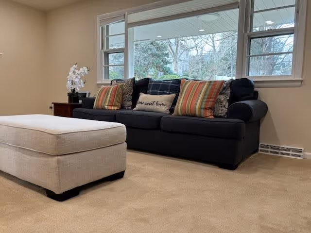 A cozy living room with a dark blue sofa adorned with multiple colorful striped and patterned cushions. In front of the sofa is a large beige ottoman. Behind the sofa, there are three large windows showing a view of trees and greenery outside. A small side table with a white orchid plant is placed next to the sofa.