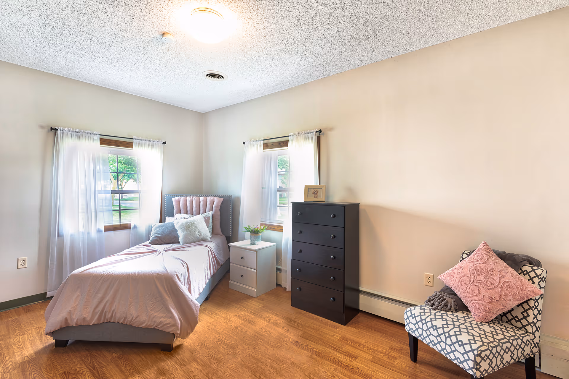 Bright single bedroom with a twin bed, two windows with sheer curtains, a nightstand, tall black dresser, and a patterned armchair.