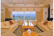 A spacious living room with two beige sofas and two beige armchairs arranged around a wooden coffee table. The room features large windows with a view of a body of water and greenery outside. The walls are decorated with light-colored curtains and ceiling fans are mounted on the ceiling.