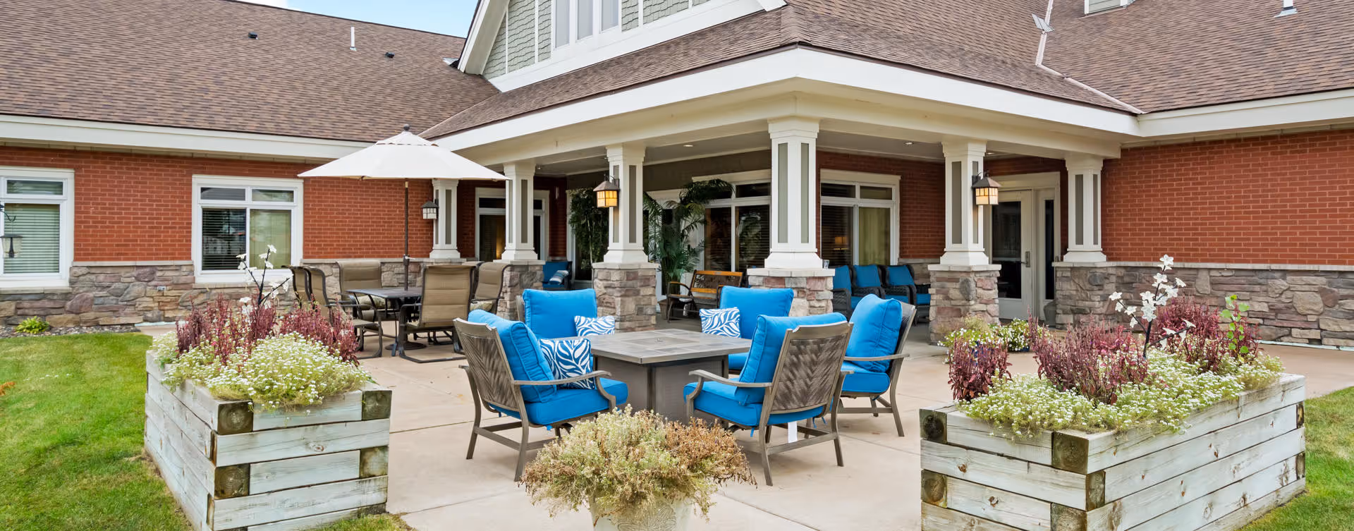 Outdoor patio area at Encore Assisted Living and Memory Care at North Branch featuring a covered seating space with blue cushioned chairs arranged around a fire pit table, additional dining chairs and tables with umbrellas, and large wooden planters filled with flowers and greenery on a concrete patio adjacent to a brick and stone building.