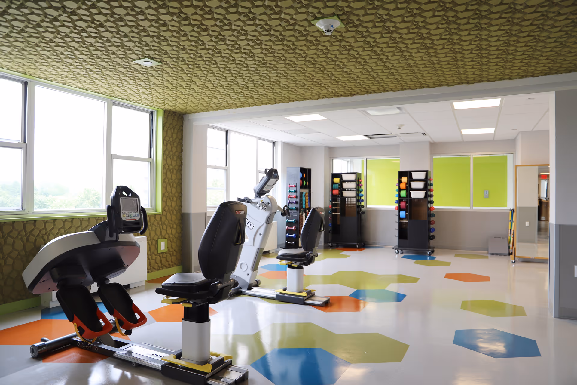 A bright exercise room with two recumbent stationary bikes, colorful hexagonal patterns on the floor, large windows letting in natural light, and racks holding various exercise balls and weights against the far wall.