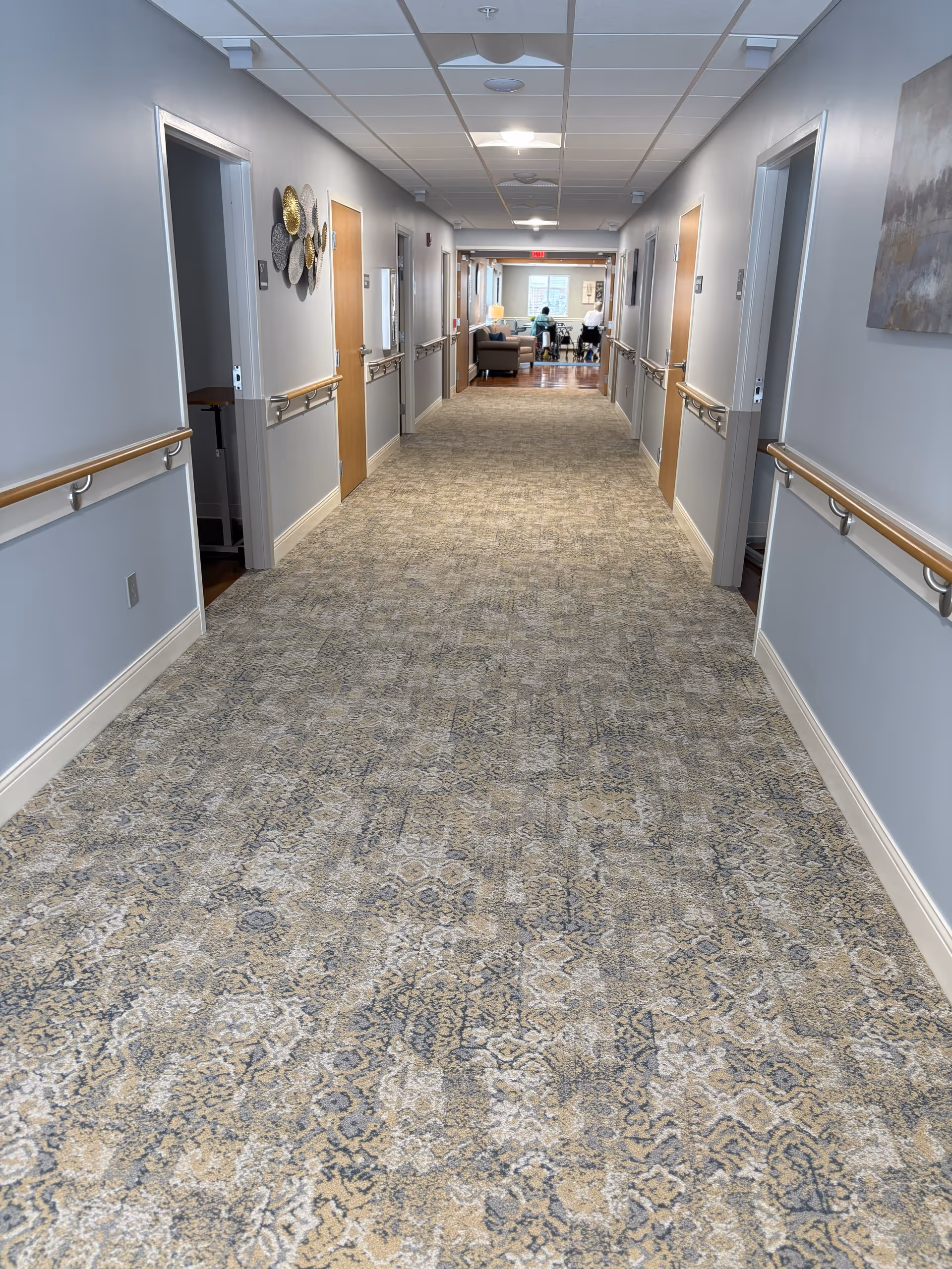 Long carpeted interior hallway with handrails and doorways leading to rooms and a seating area at the far end.