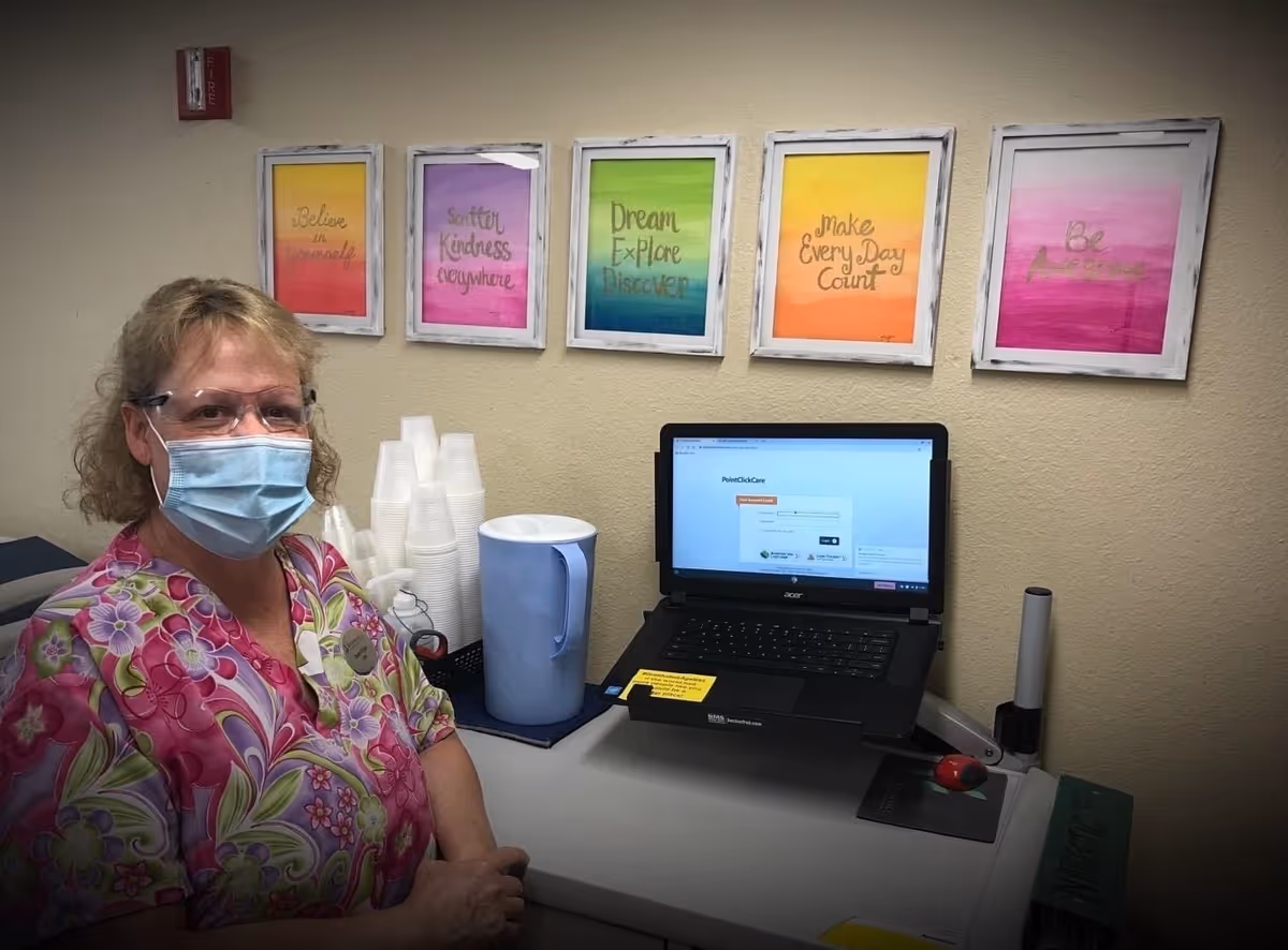 A woman wearing a floral patterned scrub top and a face mask stands next to a counter with a laptop, a blue pitcher, and stacked plastic cups. Behind her on the wall are five colorful framed motivational quotes.