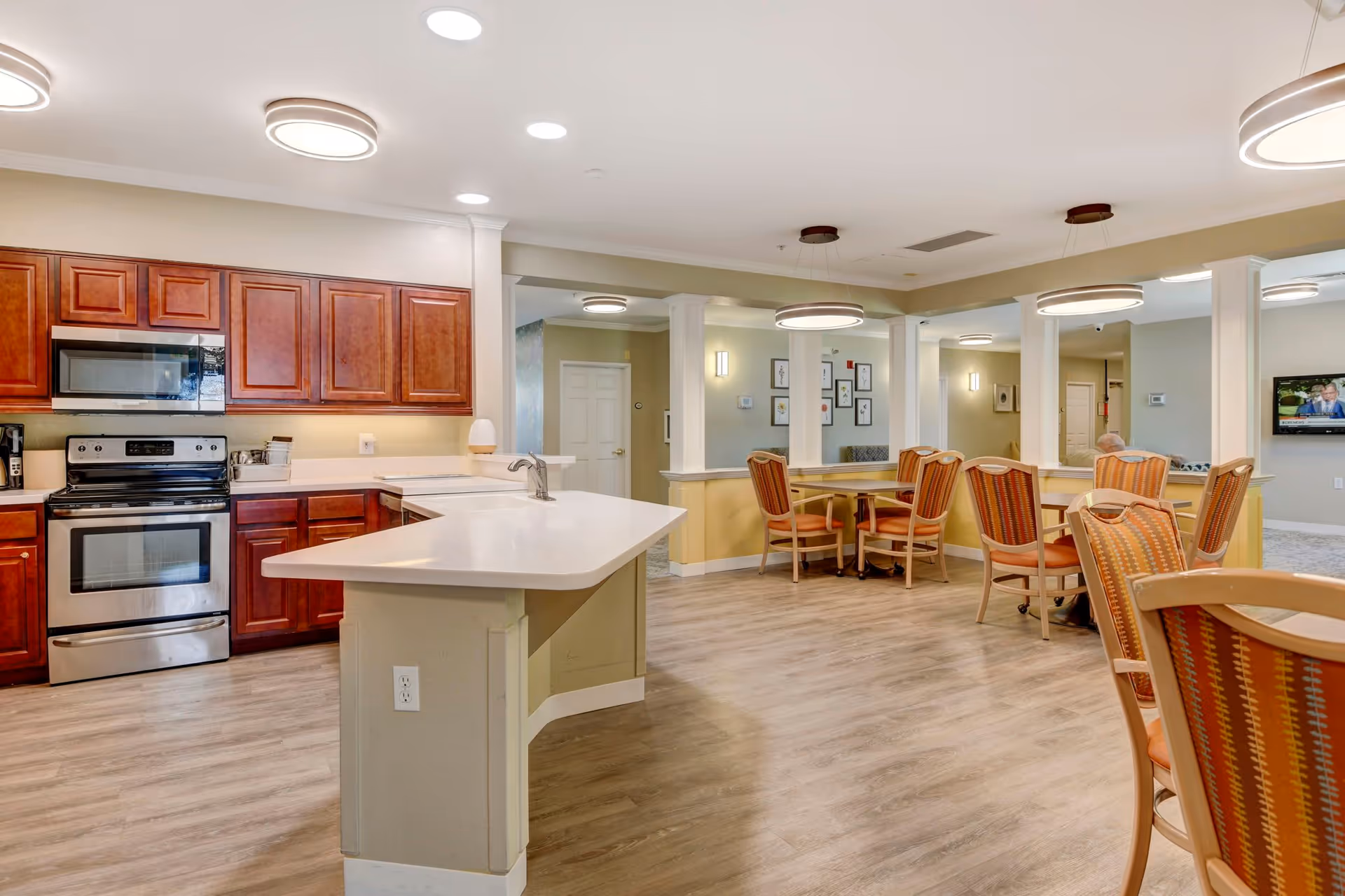 Interior view of a senior living facility showing a kitchen area with wooden cabinets, a stove, microwave, and a countertop with a sink. Adjacent to the kitchen is a dining area with multiple tables and chairs, modern circular ceiling lights, and a TV mounted on the wall in the background.