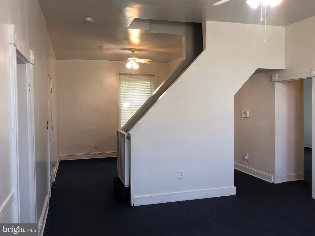 Interior view of a room with white walls and dark carpet flooring, featuring a staircase with a white railing, a ceiling fan with lights, a window with blinds, and doorways leading to other rooms.