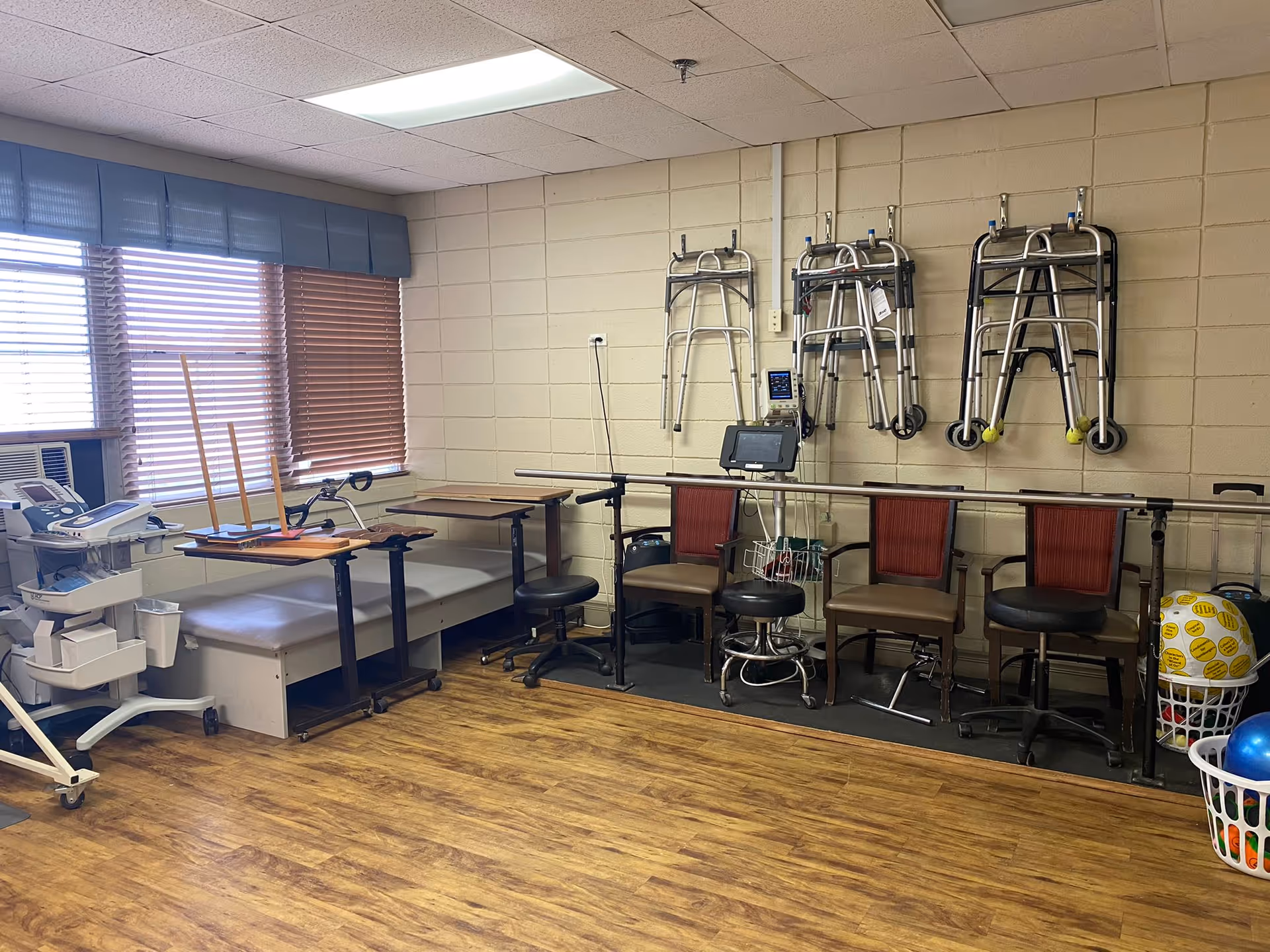 A rehabilitation or therapy room with wooden flooring, several chairs and stools, parallel bars for walking exercises, walkers hanging on the wall, and medical equipment near a window with blinds.