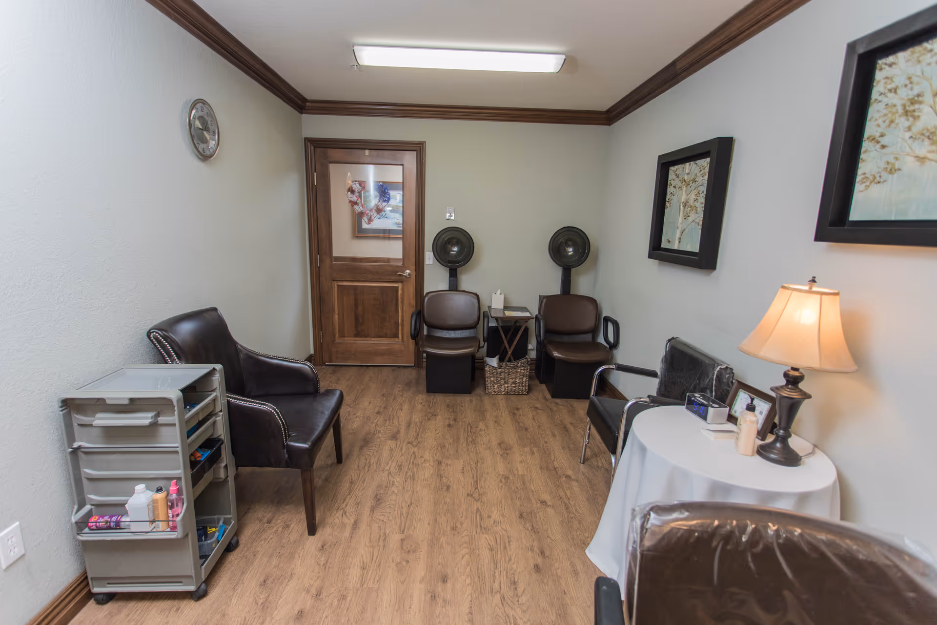 A small room with wooden flooring and beige walls featuring a wooden door at the back. The room contains several chairs, including two brown salon chairs under hair dryers, a black leather chair, and a chair with a plastic cover. There is a small round table with a lamp, lotion, and other items on it. A gray rolling cart with various supplies is on the left side. Two framed pictures hang on the walls, and a clock is mounted on the left wall.