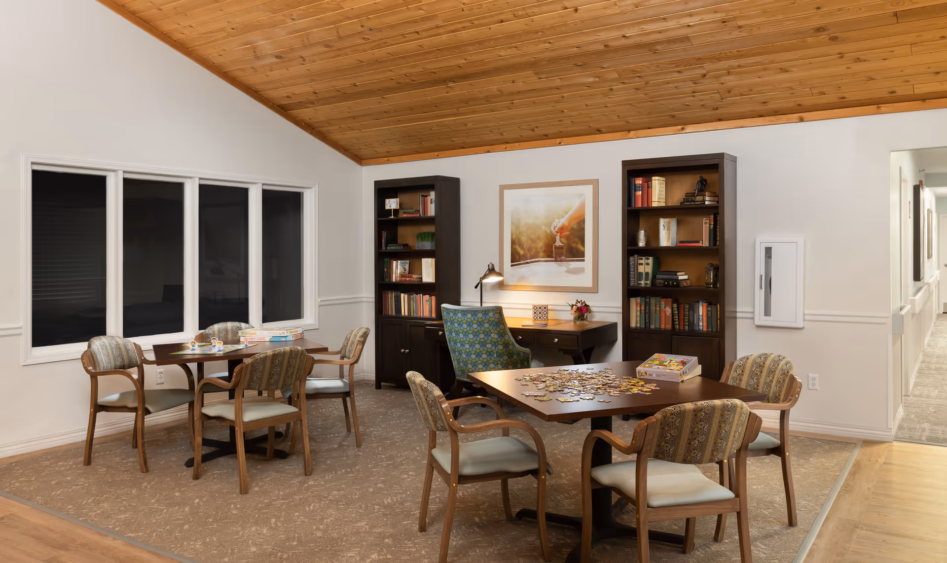 A cozy common area with two wooden tables surrounded by cushioned chairs. One table has a partially completed jigsaw puzzle and a puzzle box on it. The room features a wooden ceiling, two tall bookshelves filled with books, a desk with a lamp and a framed picture above it, and a hallway visible to the right.