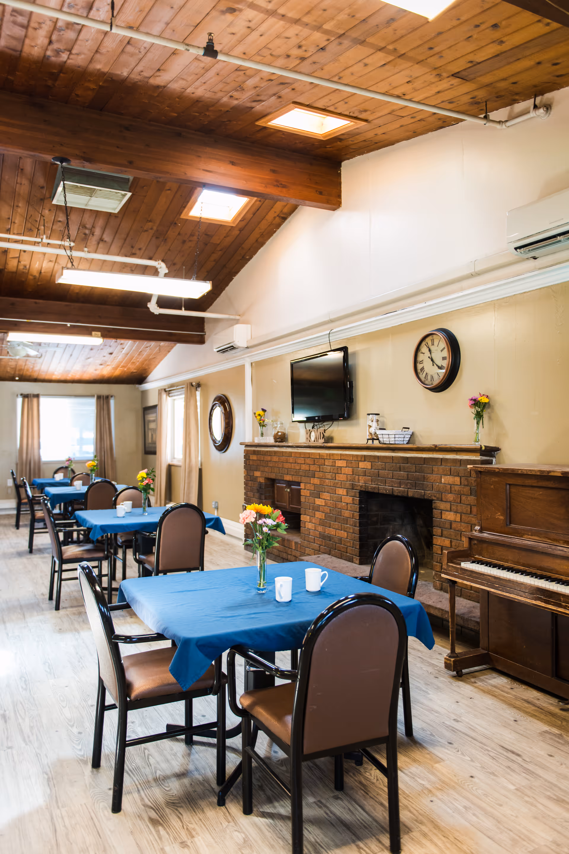 A dining room with several tables covered in blue tablecloths, each with a vase of flowers and cups. The room features a wooden ceiling with skylights, a brick fireplace with a clock and TV mounted above it, and a piano to the right. Light wood flooring and beige walls complete the cozy atmosphere.