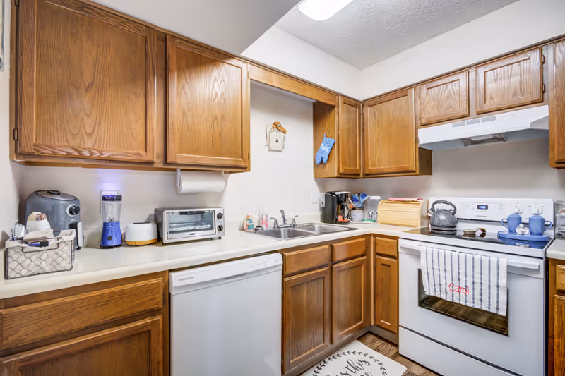 A compact kitchen with wooden cabinets, a white dishwasher, a white stove with a tea kettle on it, a toaster oven, a blender, and a coffee maker. There is a double sink with a soap dispenser and a dish brush. A striped towel with the name 'Carol' hangs on the oven door, and a blue oven mitt hangs on the wall. The countertop has various kitchen items, and the floor has a small rug.