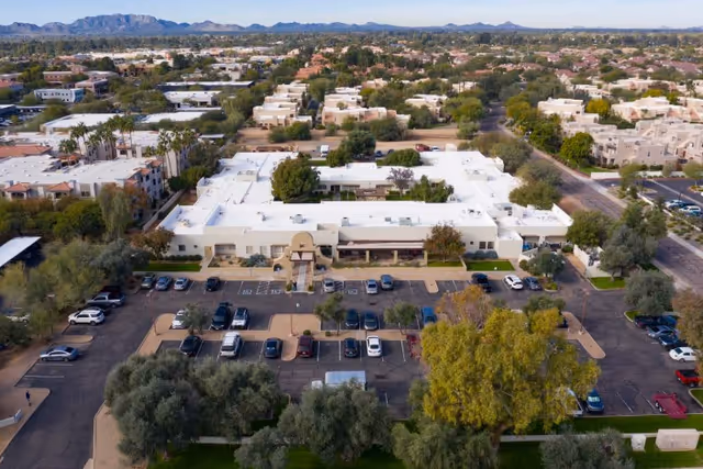 Aerial view of Shea Post Acute Rehabilitation Center showing a large single-story building with a white roof surrounded by parking lots, trees, and nearby residential areas. Mountains are visible in the background under a clear sky.