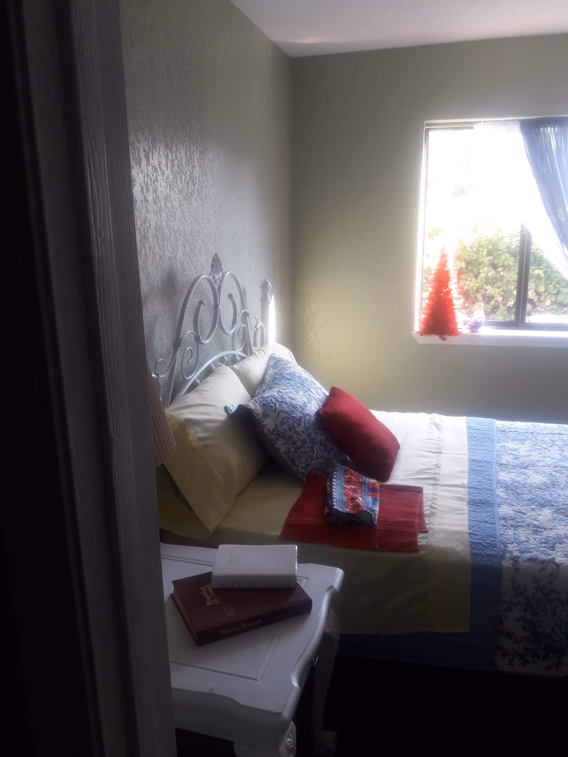 Small sunlit bedroom with a decorative metal headboard, patterned bedding, pillows and folded linens on the bed beside a white nightstand with books.