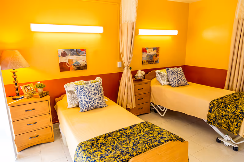 A brightly lit room with two single beds separated by a curtain. Each bed has a yellow bedspread, decorative pillows, and a patterned throw blanket. There are two wooden nightstands, one with a lamp, a framed photo, and a small plant, and the other with decorative items. The walls are painted yellow with a reddish-brown lower section, and each bed has a colorful abstract painting above it.