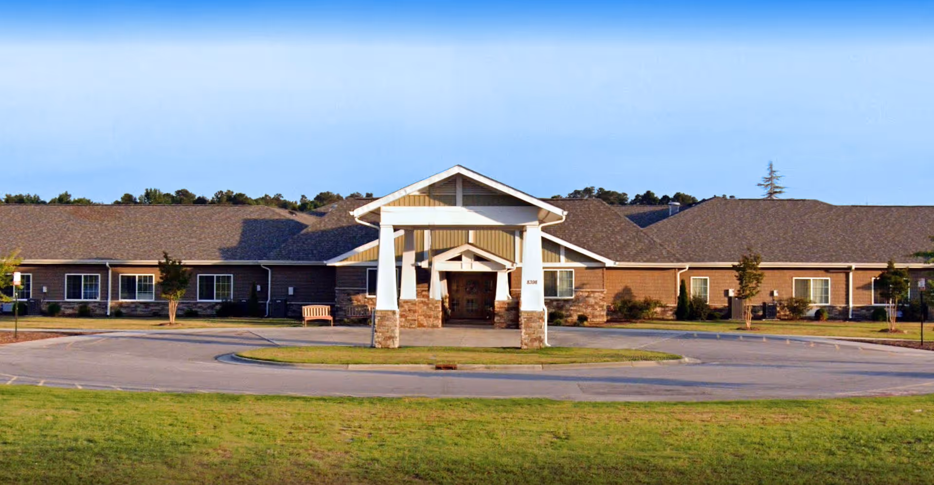 Front exterior view of a single-story senior living facility building with a covered entrance supported by white columns and a circular driveway. The building has a brown roof and beige siding with stone accents, surrounded by a grassy lawn and a few small trees.