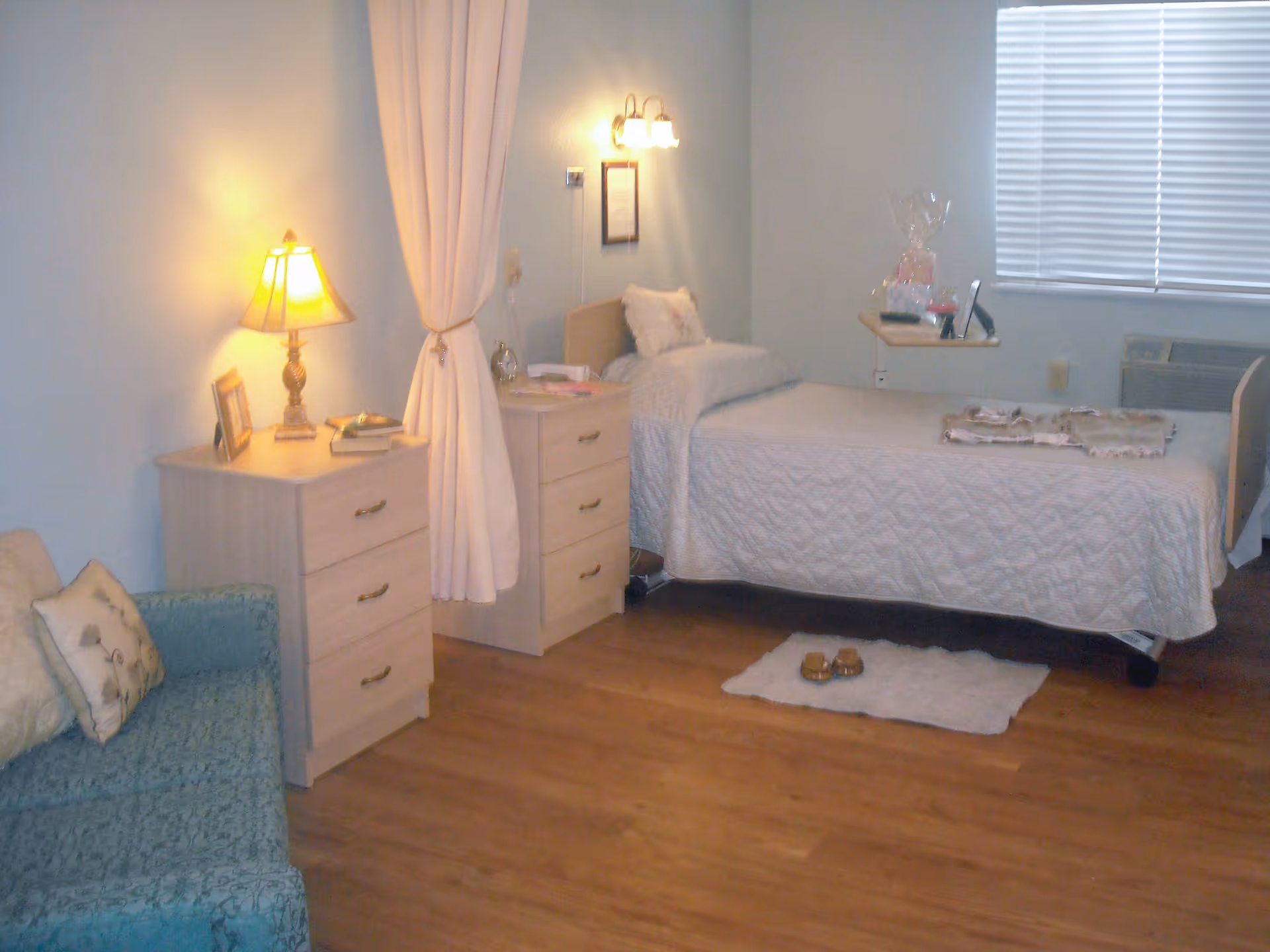 A senior living facility bedroom with a single bed covered in a white quilt, a small bedside table with a lamp, a blue patterned couch with pillows, a wooden dresser, and a window with closed blinds. The room has wooden flooring and soft lighting.