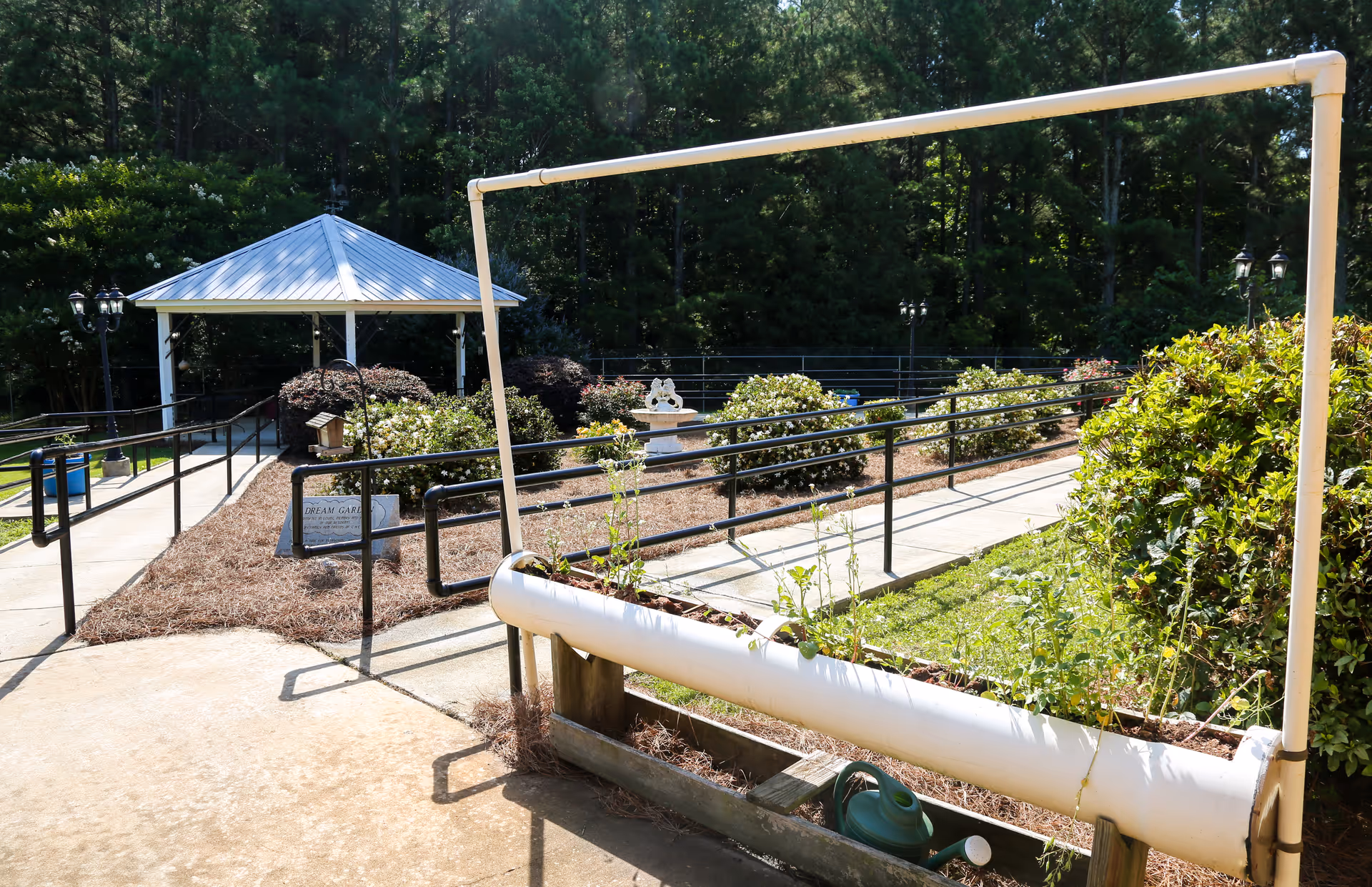 Outdoor courtyard with paved ramps, black handrails, a gazebo, and a raised PVC planter with plants.