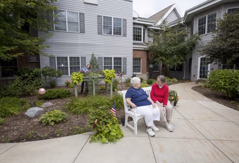 Two elderly women sitting and talking on a white bench in a garden courtyard area of a senior living facility. The courtyard is surrounded by buildings with windows and has various plants and shrubs. Another person is seen tending to plants in the background.