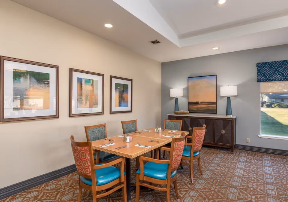 A dining room with a wooden table set for six people with utensils and napkins. The chairs have wooden frames with teal and patterned upholstery. On the wall to the left are three framed abstract art pieces, and on the far wall is a sideboard with two lamps and a large framed painting above it. A window with a blue patterned valance lets in natural light, showing a grassy outdoor area.