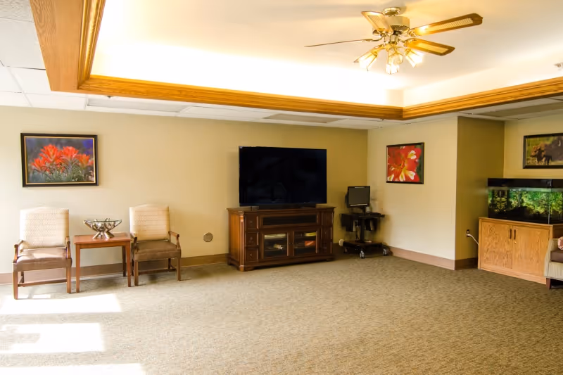 A well-lit common area with beige walls and carpeted floor. The room features a ceiling fan with lights, two chairs with a small table between them, a large flat-screen TV on a wooden cabinet, a small computer workstation on wheels, and a large fish tank on a wooden stand. There are three framed pictures on the walls.