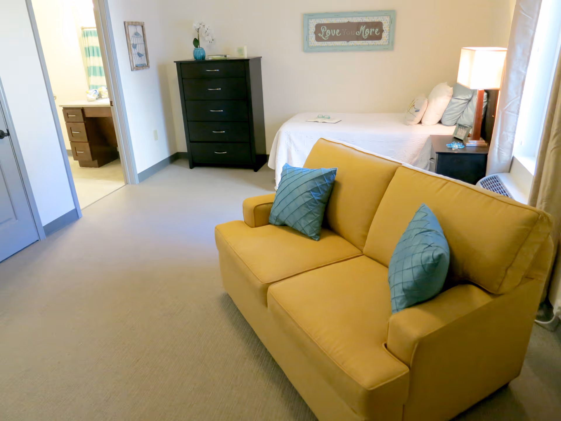 A cozy assisted living room featuring a yellow loveseat with two blue pillows, a single bed with white bedding and multiple pillows, a black nightstand with a lamp, a black chest of drawers, and a bathroom visible through an open door. A framed sign above the bed reads 'Love You More'.