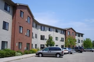Exterior view of a multi-story senior living facility building with a combination of brick and light-colored siding. Several cars are parked in front of the building under a clear blue sky.
