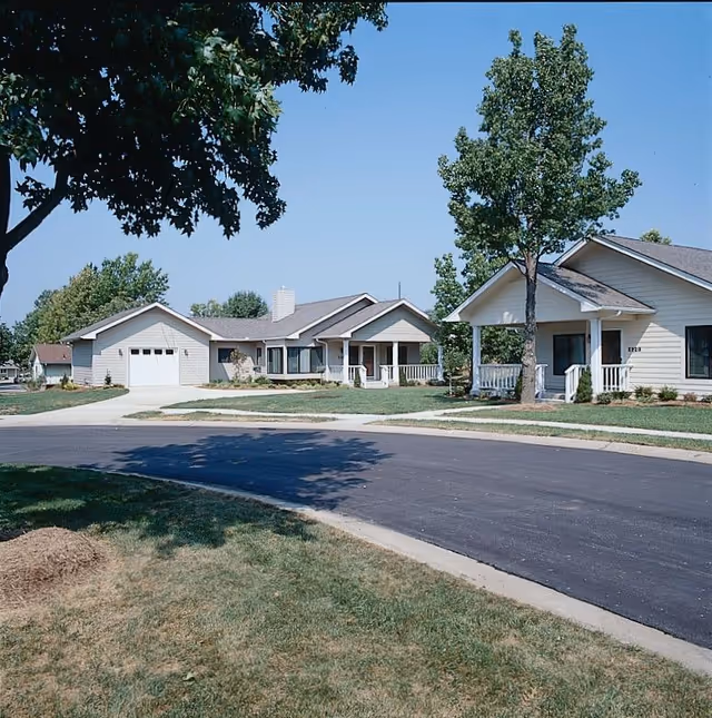 A quiet residential street in a senior living community with single-story houses featuring porches, garages, and well-maintained lawns under a clear blue sky.