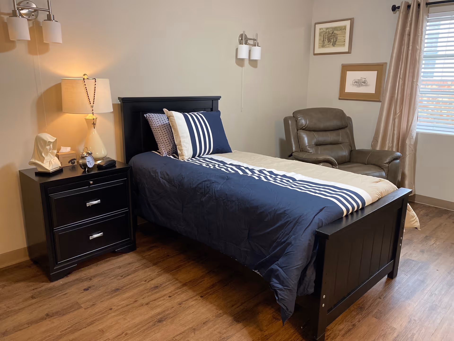 A neatly made single bed with navy blue and white striped bedding in a senior living bedroom. Next to the bed is a black nightstand with a lamp, a clock, a tissue box, and a white bust sculpture. A brown leather recliner chair is positioned near a window with beige curtains. Two framed pictures hang on the wall above the chair. The room has wooden flooring and soft lighting.