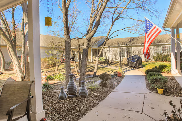 Covered courtyard walkway with trees, decorative planters, chairs, and an American flag in front of a single-story assisted living building.