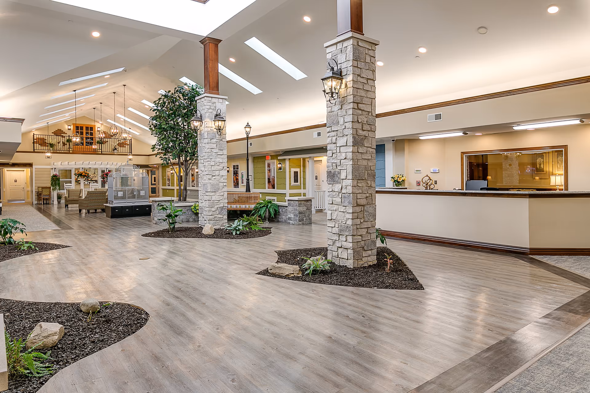 Spacious and well-lit interior common area of an assisted living facility featuring stone pillars with mounted lantern-style lights, indoor plants, a birdcage, seating areas with chairs and benches, and a reception desk on the right side. The ceiling has skylights and hanging light fixtures, and the floor is covered with light wood-style flooring.