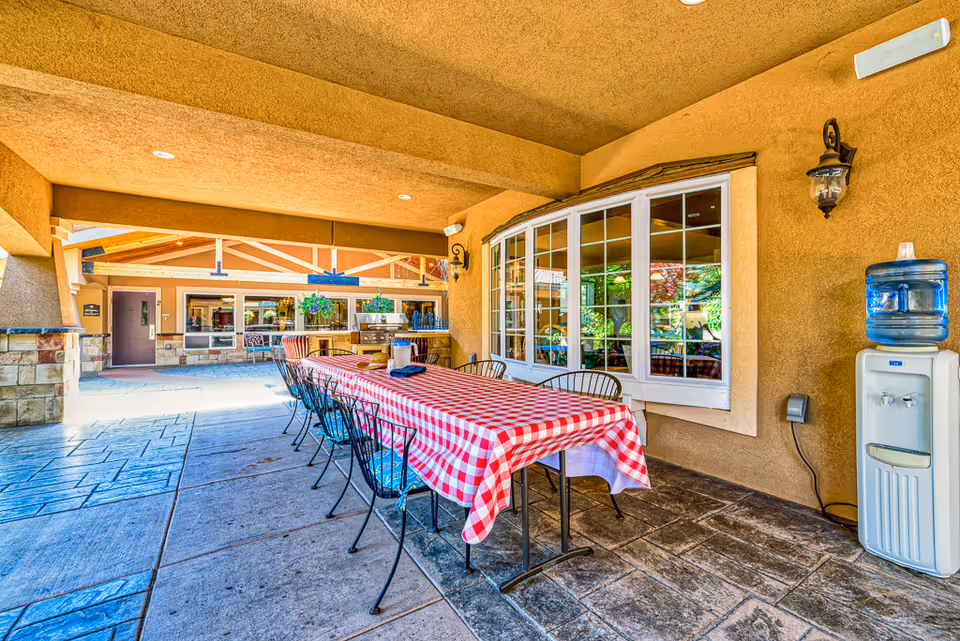Covered outdoor patio area with a long table covered in a red and white checkered tablecloth surrounded by metal chairs. There is a water cooler on the right side and large windows looking into an interior space. The patio has stone flooring and beige stucco walls with decorative lighting.