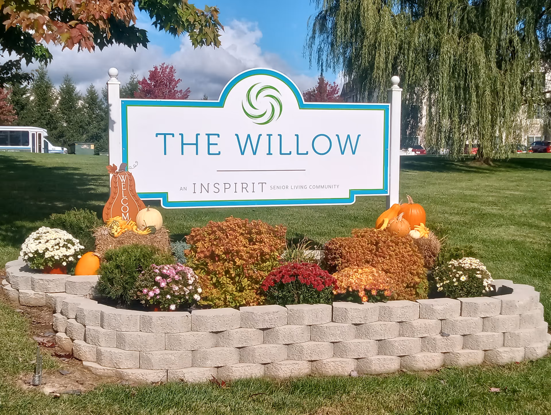 Landscaped entrance sign reading "The Willow" with pumpkins and fall flowers on a grassy lawn.